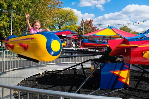A young girl in a pink shirt waves from a colorful amusement park ride under a blue sky with fluffy clouds.