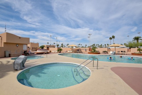 A sunny pool area featuring a large swimming pool, a hot tub, and palm trees under a blue sky.