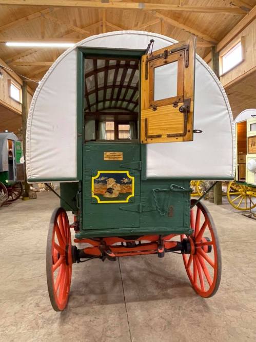 A vintage green and white covered wagon with red wheels, featuring a wooden door and decorative emblem.
