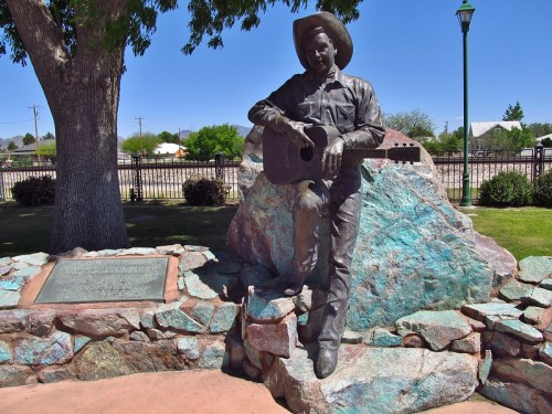 Bronze statue of a cowboy with a guitar, seated on a rock in a park, surrounded by greenery and a stone wall.
