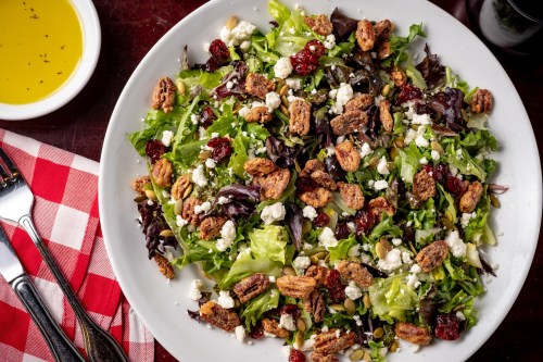 A vibrant salad with mixed greens, cranberries, pecans, feta cheese, and a side of olive oil on a checkered tablecloth.