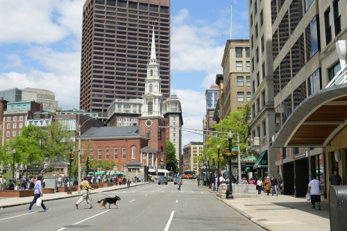 A busy city street with pedestrians, a dog, and historic buildings under a partly cloudy sky.