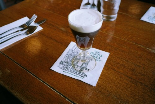 A glass of coffee topped with foam sits on a napkin featuring a city illustration, on a wooden table.