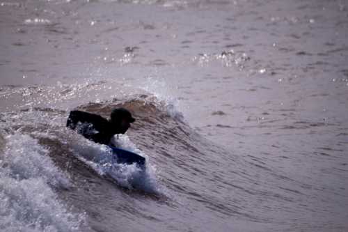 A person in a wetsuit rides a small wave on a bodyboard, creating splashes in the water.