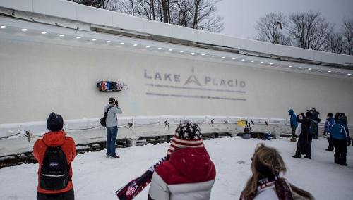 A bobsledder races down an icy track in Lake Placid, while spectators watch from the sidelines in snowy conditions.