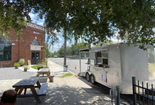 A food truck parked near a brick building with outdoor seating under trees on a sunny day.