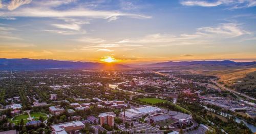Aerial view of a city at sunset, with mountains in the background and a river winding through green parks and buildings.