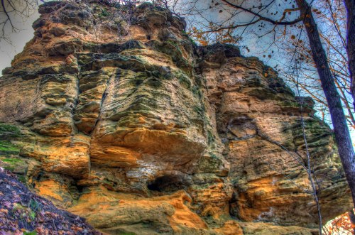 A rugged rock formation with layered textures and colors, surrounded by trees and foliage.