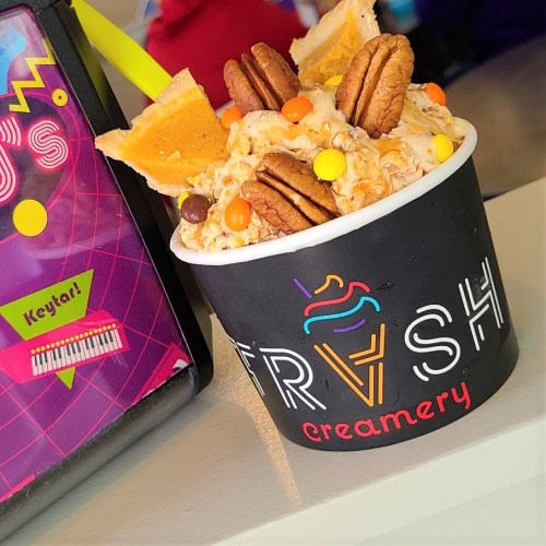 A colorful ice cream bowl topped with pecans, candy, and waffle pieces, placed next to a vibrant menu display.