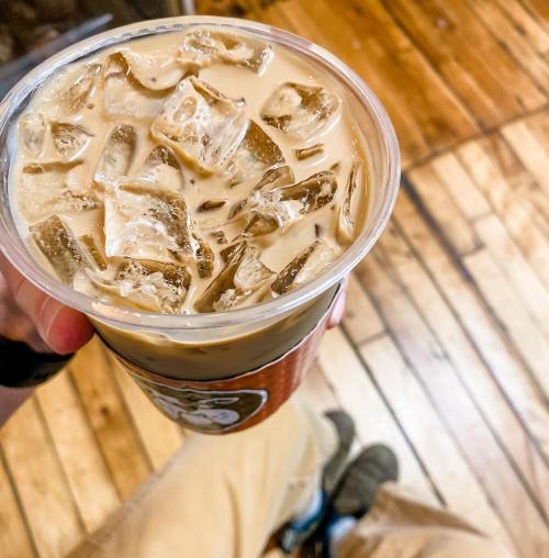 A close-up of a hand holding a cup of iced coffee with ice cubes, set against a wooden floor background.