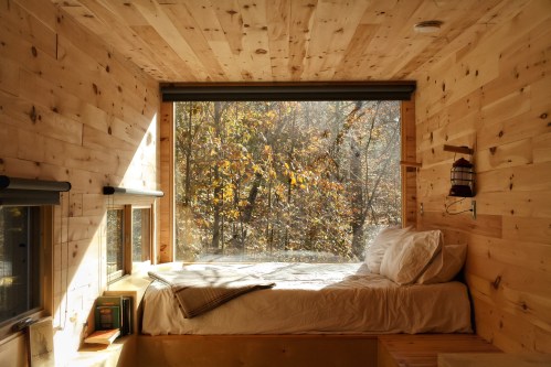 Cozy wooden bedroom with a large window overlooking autumn trees, featuring a bed and books on a shelf.