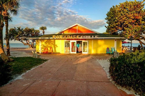 A bright yellow beachside restaurant named "Paradise Grille," surrounded by palm trees and sandy pathways.