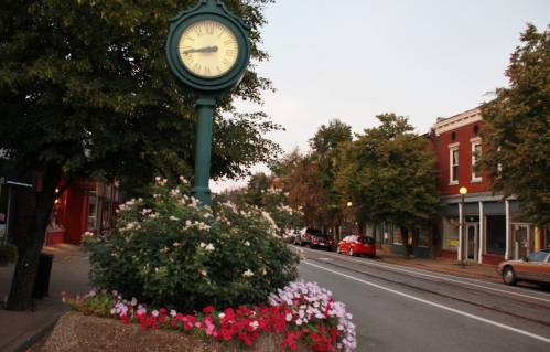 A vintage clock stands in a flower bed along a quiet street lined with trees and charming buildings at dusk.