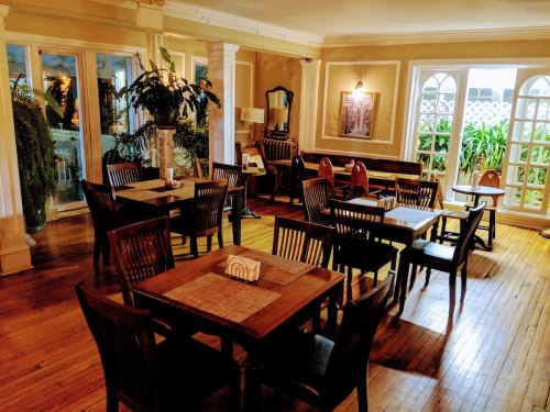 Cozy restaurant interior with wooden tables, chairs, and plants, featuring large windows and warm lighting.