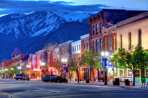A charming street lined with colorful buildings and trees, set against a backdrop of snow-capped mountains at dusk.