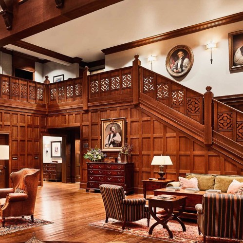 A cozy, wooden-paneled interior with a staircase, elegant furniture, and framed portraits on the walls.