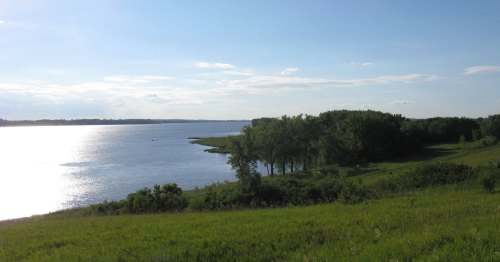 A serene landscape featuring a calm river, lush green banks, and a clear blue sky with scattered clouds.