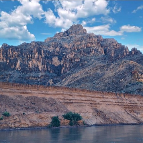 A rugged mountain rises above a riverbank, with dramatic cliffs and a blue sky dotted with clouds.