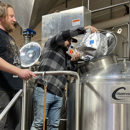 Two men work together in a brewery, one pouring ingredients into a large stainless steel tank.
