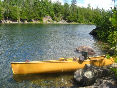A yellow canoe rests on a rocky shore by a calm, clear lake surrounded by lush green trees.