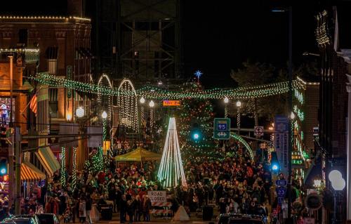 A festive street scene at night, filled with people, holiday lights, and a large Christmas tree.