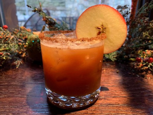 A cocktail with a cinnamon-sugar rim, garnished with an apple slice, sits on a wooden surface surrounded by greenery.