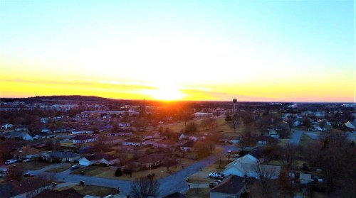 Aerial view of a suburban neighborhood at sunset, with vibrant orange and blue skies and scattered houses below.