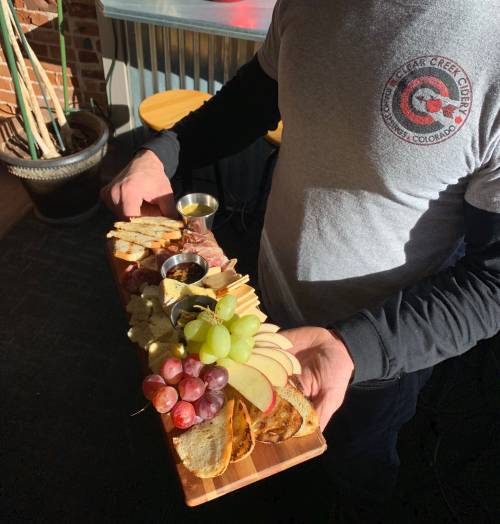 A person holds a wooden platter with assorted cheeses, meats, fruits, and bread, accompanied by small dipping sauces.