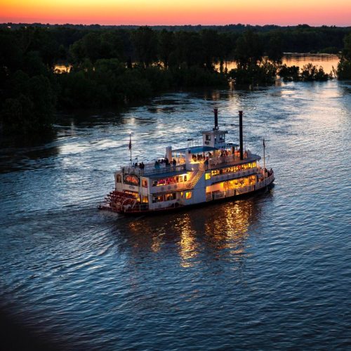 A riverboat glides through calm waters at sunset, reflecting warm colors and surrounded by lush greenery.
