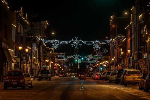 A festive downtown street at night, adorned with snowflake lights and lined with parked cars.
