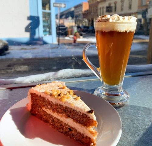 A slice of cake on a plate next to a glass of coffee topped with cream, with a sunny street view in the background.