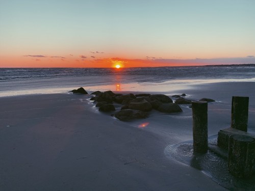 A serene beach at sunset, with the sun setting over the horizon and rocks scattered along the shore.