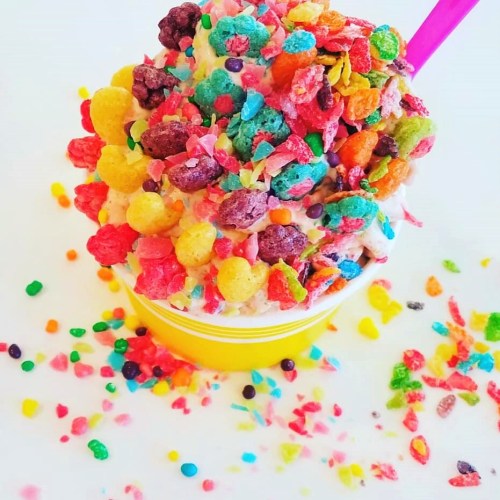 A colorful ice cream topped with vibrant cereal and surrounded by scattered cereal pieces on a white surface.