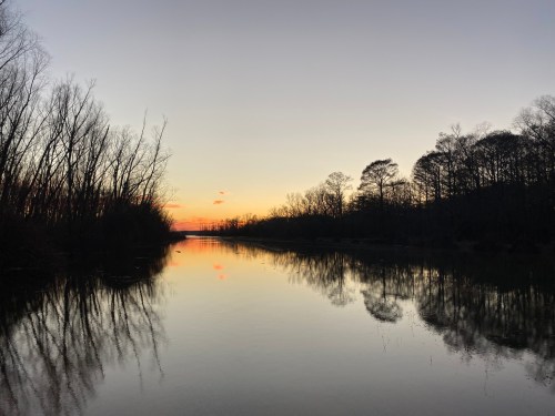 A serene river at sunset, with trees lining the banks and reflections on the calm water.