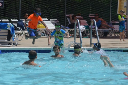 Children playing in a pool, some jumping in while others swim, with adults relaxing on lounge chairs nearby.