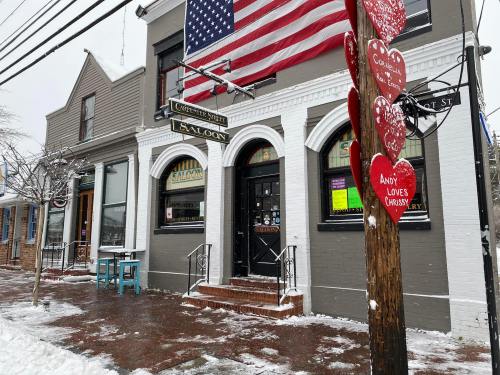 A snowy street scene featuring a saloon with an American flag and heart decorations on a nearby pole.