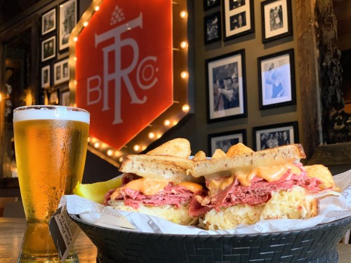 A close-up of a hearty sandwich with layers of meat and cheese, accompanied by a glass of beer, set against a cozy backdrop.