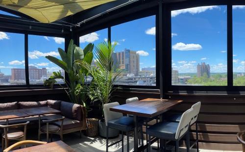 A rooftop dining area with large windows, plants, and wooden tables, overlooking a cityscape under a blue sky.