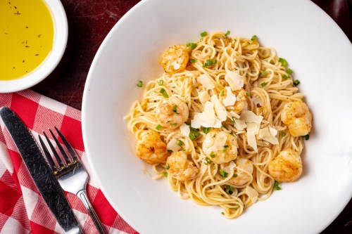 A bowl of shrimp pasta topped with parmesan and herbs, served with olive oil on the side and a checkered napkin.