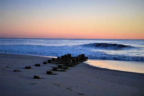 A serene beach at sunset, featuring wooden posts leading into gentle waves and a colorful sky.