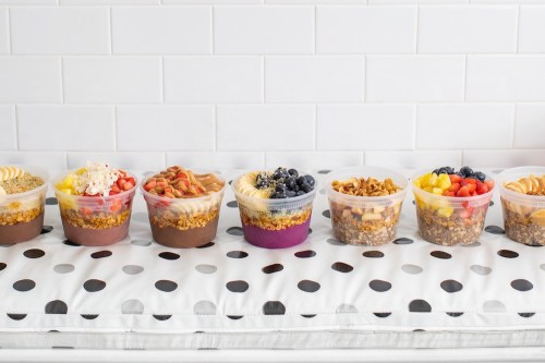 Seven colorful dessert cups filled with layers of yogurt, granola, fruits, and toppings, arranged on a polka dot surface.