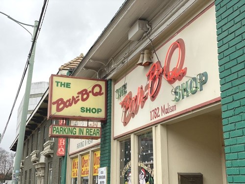 Signage for "The Bar-B-Q Shop" with colorful lettering, featuring a parking notice, on a brick building.