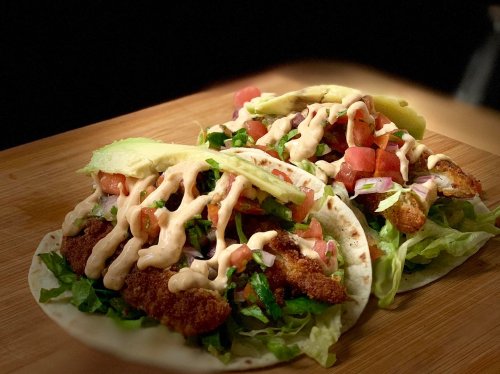 Two tacos filled with lettuce, tomatoes, and crispy fish, drizzled with a creamy sauce, on a wooden surface.
