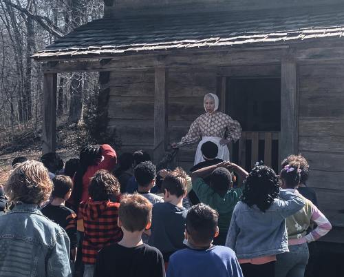 A historical reenactor speaks to a group of children outside a rustic cabin in a wooded area.