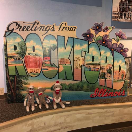 A colorful "Greetings from Rockford, Illinois" sign with three sock monkeys in front of it.