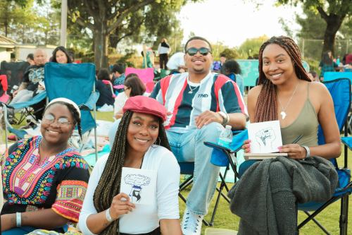 A group of four friends sitting outdoors, smiling and holding drawings, with a lively crowd in the background.