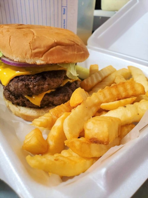 A double cheeseburger with lettuce, onion, and cheese, served with crinkle-cut fries in a takeout container.