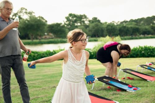 A girl in a white dress and glasses prepares to throw a bean bag while adults play cornhole by a lake.