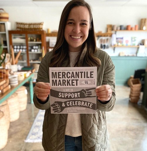 A smiling woman holds a flyer for the Mercantile Market, promoting a day to support and celebrate local craftsmanship.