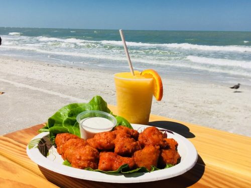 A plate of golden fried food with a dipping sauce, next to a tropical drink, set against a beach and ocean backdrop.